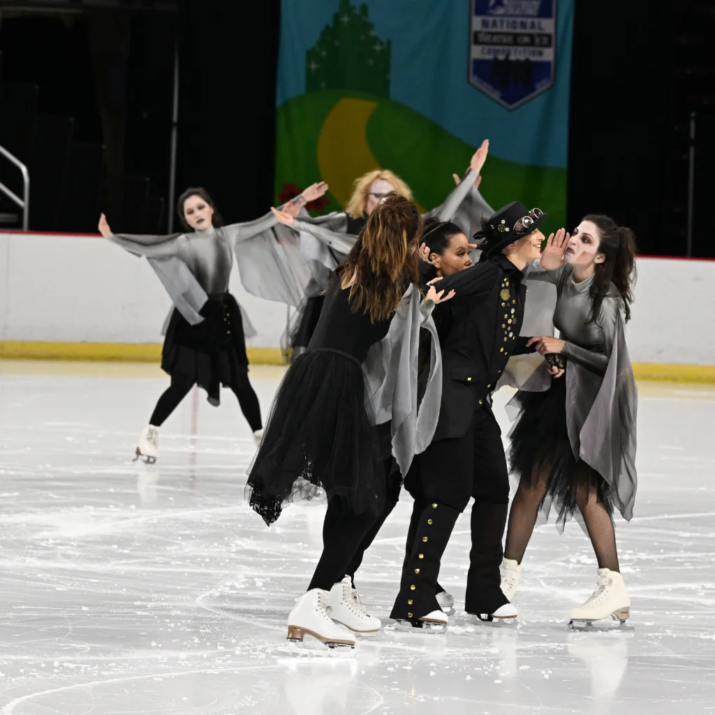 Figure skaters in grey "ghostly" costume dresses whispering into the ear of a skater in a steampunk suit and top hat