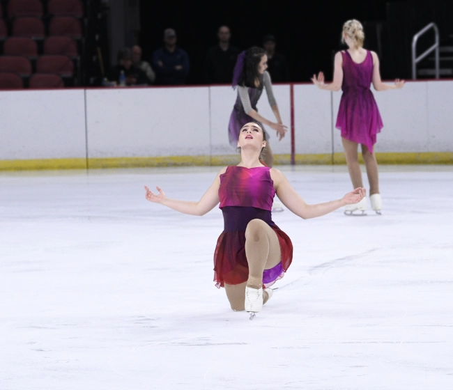 Female figure skater in pink and maroon dress with arms outstretched and looking to the sky